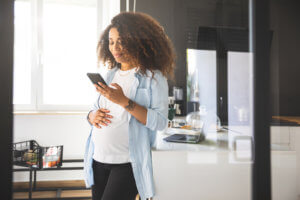 Pregnant woman with curly hair texting on mobile and scrolling social media in a bright morning in her modern and white kitchen.